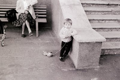 A young child in 1960s-era clothing holds a leash while a small dog sits beside a concrete stairwell. A woman in a plaid skir...