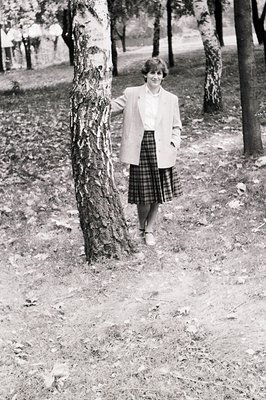 Black-and-white portrait of a woman in a wooded park, leaning against a tree trunk. She wears a white blouse, a plaid skirt, ...