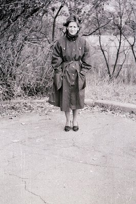 Portrait of a woman in a structured 1940s-50s coat, standing on a paved path with autumn foliage in background. Her attire in...