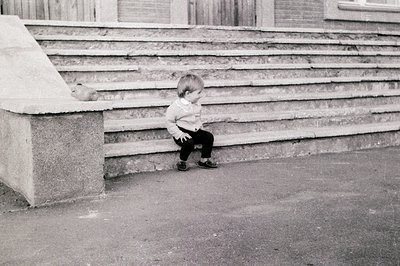 Young child sitting on concrete steps, clutching a stuffed toy, mid-1960s–70s urban setting. Monochrome photo captures candid...