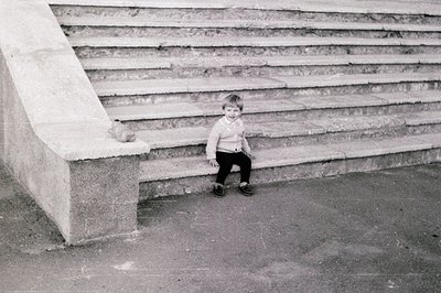 Young child sitting on concrete steps, mid-1970s urban setting. Wearing a striped sweater and dark pants, posing with a relax...