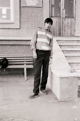 Young man in a striped sweater and dark trousers stands on concrete steps outside a brick building, 1960s-70s urban setting.