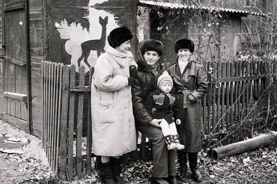 Four individuals pose outdoors in winter attire, likely Eastern Europe, mid-20th century. A mural of a deer and tree stump ad...