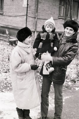 Black-and-white snapshot of three individuals outdoors in winter, likely 1970s–1980s. A woman in a long coat holds a child bu...