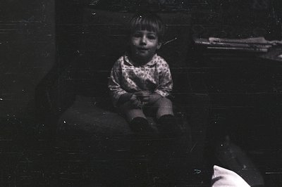 Vintage black-and-white photo of a young child sitting on a dark, textured surface, possibly a bench or ledge. The child wear...