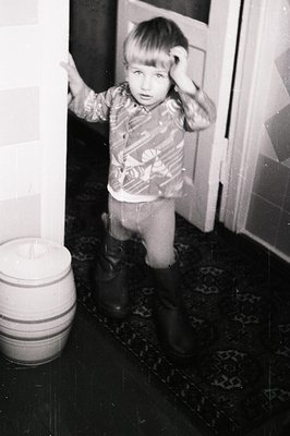 Young child in 1960s-70s indoor setting, posing with hands on head. Wears patterned sweater, knee-length pants, and dark shoe...