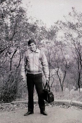 Black-and-white portrait of a man in a quilted jacket and trousers, standing on a gravel path surrounded by leafless trees, s...