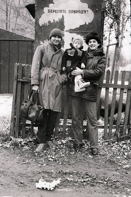 Three women pose outdoors in winter attire, holding a child and suitcases. Poster in Cyrillic reads "Берегите природу!" (Prot...