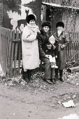 Three women in winter attire pose outdoors beside a wooden fence, holding a child. The central mural depicts a bird in flight...