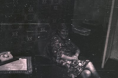 Vintage black-and-white photo of a seated woman in a patterned dress, holding a small object. Rotary dial phone and woven rug...