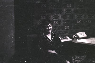Vintage black-and-white portrait of a woman seated indoors, likely mid-20th century. She wears a high-necked blouse and sits ...
