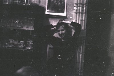 Vintage black-and-white photo of a person laughing in a dimly lit interior, likely a 1950s–1960s home. Reflections in a mirro...