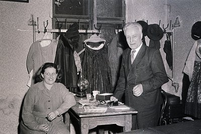 Vintage black-and-white photo of a 1940s-50s clothing shop interior. A man in a suit and woman in a patterned dress pose near...