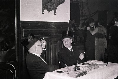 Two men in formal 1940s-50s attire—wide-lapelled suits, fedoras, and pocket watches—sit at a table with books, pens, and a de...
