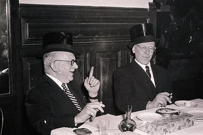 Two elderly men in formal attire—vests, ties, and fedoras—engage in conversation at a table in a dimly lit indoor setting, li...