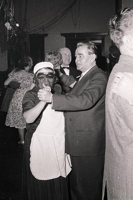 A black-and-white photo capturing a 1950s–60s formal event. A masked woman in a white apron and skirt dances with a man in a ...