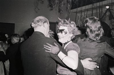 Mid-20th century indoor party scene with a woman in a horned headdress and sunglasses hugging a man in a suit. Crowded room w...