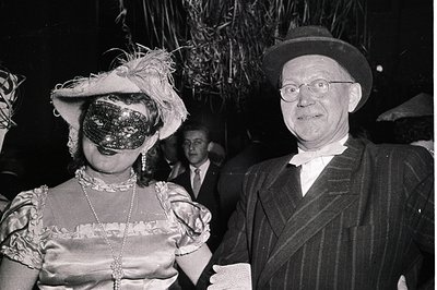Black-and-white 1930s-40s formal event photo: woman in elaborate 1920s-style dress with feathered hat and decorative mask, ma...
