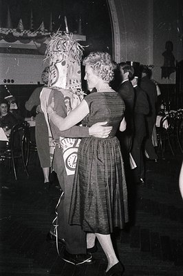 Mid-20th century indoor dance event featuring a woman in a 1950s-style dress embracing a performer in a detailed, feathered h...