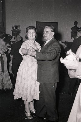 Couple dancing in a formal indoor setting, likely a 1950s–1960s social event. Woman in a fitted floral dress, man in a suit. ...