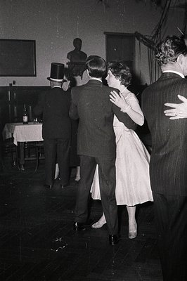 Mid-20th century social gathering in a formal indoor setting, likely 1950s–1960s. Couple dancing in a ballroom-style venue, w...