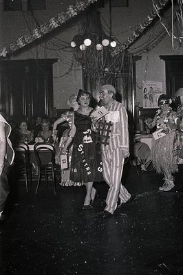 Black-and-white photo of a 1960s/70s costume party in a dimly lit indoor venue. Central figures wear playful outfits: a woman...