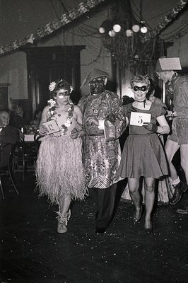 Three women in 1940s-50s masquerade costumes—tiki skirt, ornate mask, and vintage dress—walk indoors at a festive event. Deco...
