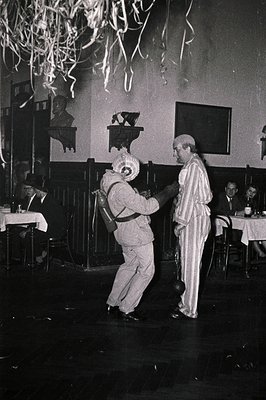 Black-and-white photo captures a 1950s–60s indoor celebration: two dancers in formalwear (man in light trousers, woman in str...