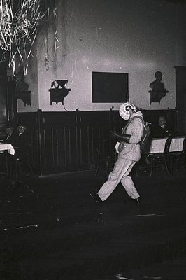 Black-and-white photo of a person in a white mask and 1970s-style ensemble dancing in a dimly lit hall. Wooden benches, frame...