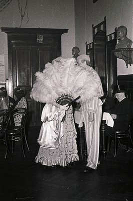 Black-and-white formal event featuring two women in elaborate 19th-century attire: one in a voluminous lace gown with a feath...