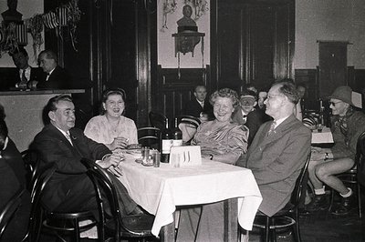 Mid-century indoor gathering in a formal dining setting, featuring six adults seated around a table with vintage tableware. M...