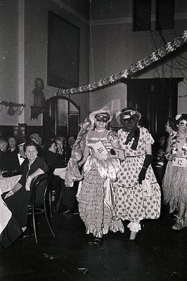 Vintage black-and-white photo of a 1950s-60s costume party. Two women in elaborate, retro gowns—one with a ruffled skirt and ...