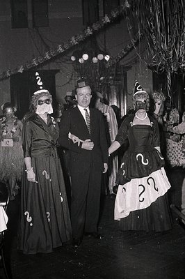 Three individuals in 1950s-style question-mark costumes pose indoors, likely at a party or event. The man wears a suit with a...