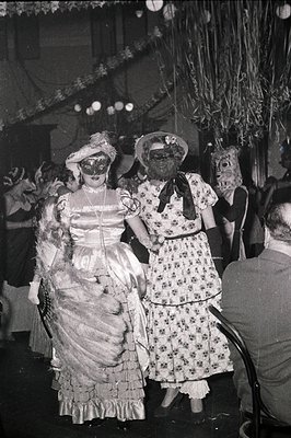 Vintage black-and-white photo of two women in elaborate 1920s-30s masquerade costumes: one in a feathered, draped gown with a...