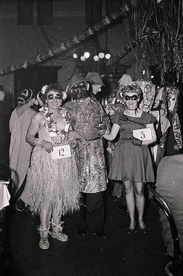 Black-and-white photo of three costumed participants at a 1950s–60s masquerade event. Left: Woman in grass skirt, floral head...