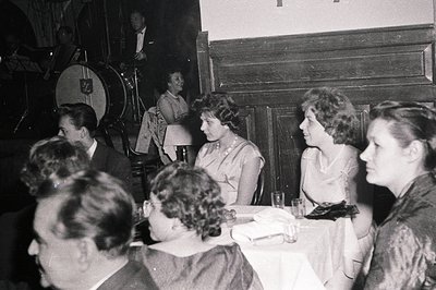 Vintage black-and-white photo of a mid-20th-century social gathering in a dimly lit room. A group of men and women seated at ...