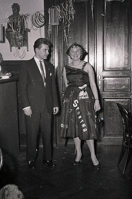 Black-and-white 1960s formal portrait: man in dark suit with lapel pin, woman in knee-length dress with "SPORTHA" appliqué. I...