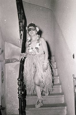Mid-20th century black-and-white photo of a person in a grass skirt and floral lei posing on ornate wrought-iron staircase. C...
