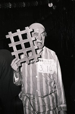 Prisoner-of-war striped uniform with chain belt and numbered tag "3.14" over heart, holding a barbed-wire cross prop. Likely ...