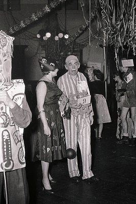 Vintage black-and-white photo of a 1950s-60s costume party. Central figure wears a clown outfit with striped pants, suspender...