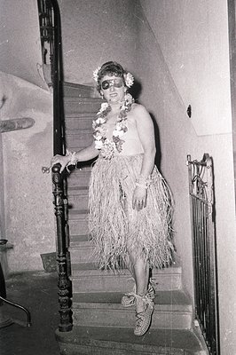 Mid-century tropical-inspired attire featuring a grass skirt, floral headband, and sunglasses. Indoor setting with ornate wro...