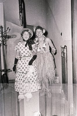 Vintage black-and-white photo of two women in 1930s-40s Hawaiian-themed costumes, posing indoors. Left: floral-patterned dres...