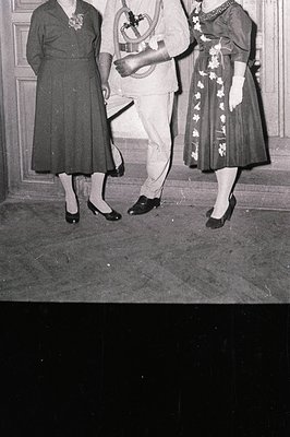 Black-and-white photo capturing mid-20th century formal attire—women in knee-length dresses with floral embroidery and brooch...