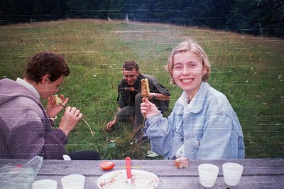 Vintage outdoor picnic scene with three individuals enjoying food by a rustic wooden table. The woman in a denim jacket holds...