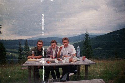 Three individuals seated at a rustic wooden picnic table in a mountainous landscape, likely . The scene captures a casual out...