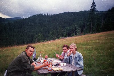 Vintage outdoor picnic scene with three individuals seated at a wooden table on a grassy hillside. Forest and rolling terrain...