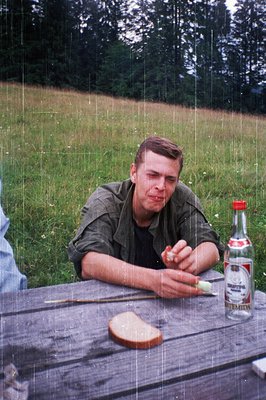 Man in 1990s outdoor setting, seated at a wooden picnic table under light rain. He wears a green jacket, holds a lit cigarett...
