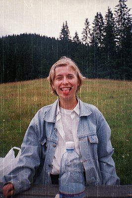 A woman in a light blue denim jacket and white blouse stands in light rain, smiling under a forest backdrop. She holds a plas...