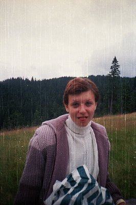 Portrait of a person in a light sweater and cardigan standing in light rain, with a forested hillside and lone tree in backgr...