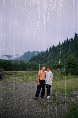 Two men pose under a rain curtain in a rural, forested landscape. The vertical water streams create a dynamic, artistic backd...